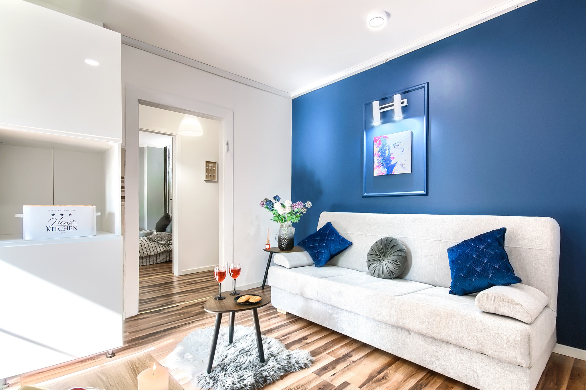 Jasny salon z akcentem granatowej ściany, białym narożnikiem z poduszkami, stolikiem z przekąskami i widokiem na sypialnię w tle.