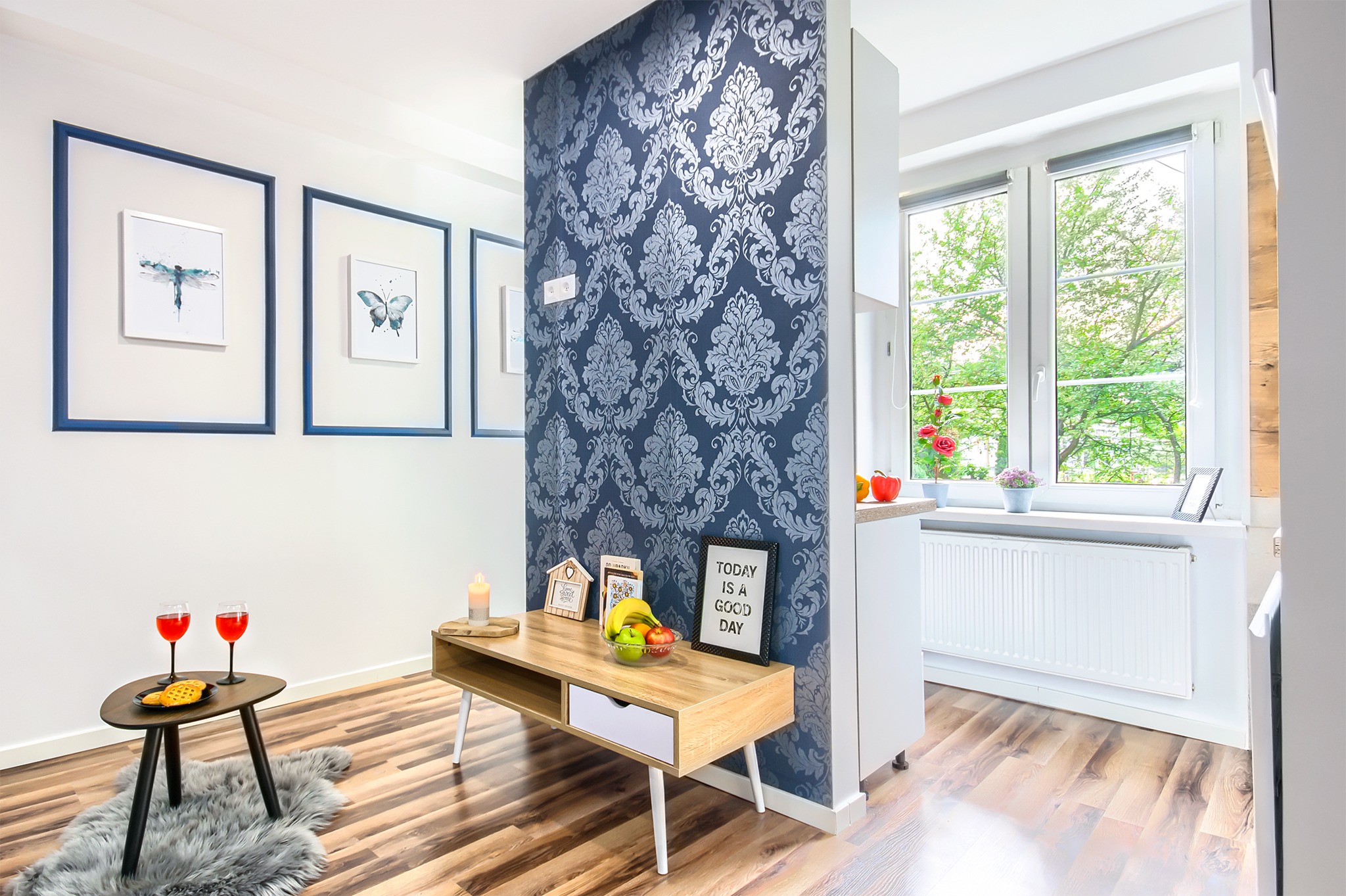 Jasne wnętrze kawalerki po liftingu w Jeleniej Górze: drewniana podłoga, stolik z napojami i przekąskami na futrzanym dywaniku, dekoracyjna ściana z wzorzystą tapetą, trzy obrazy z motywami owadów...