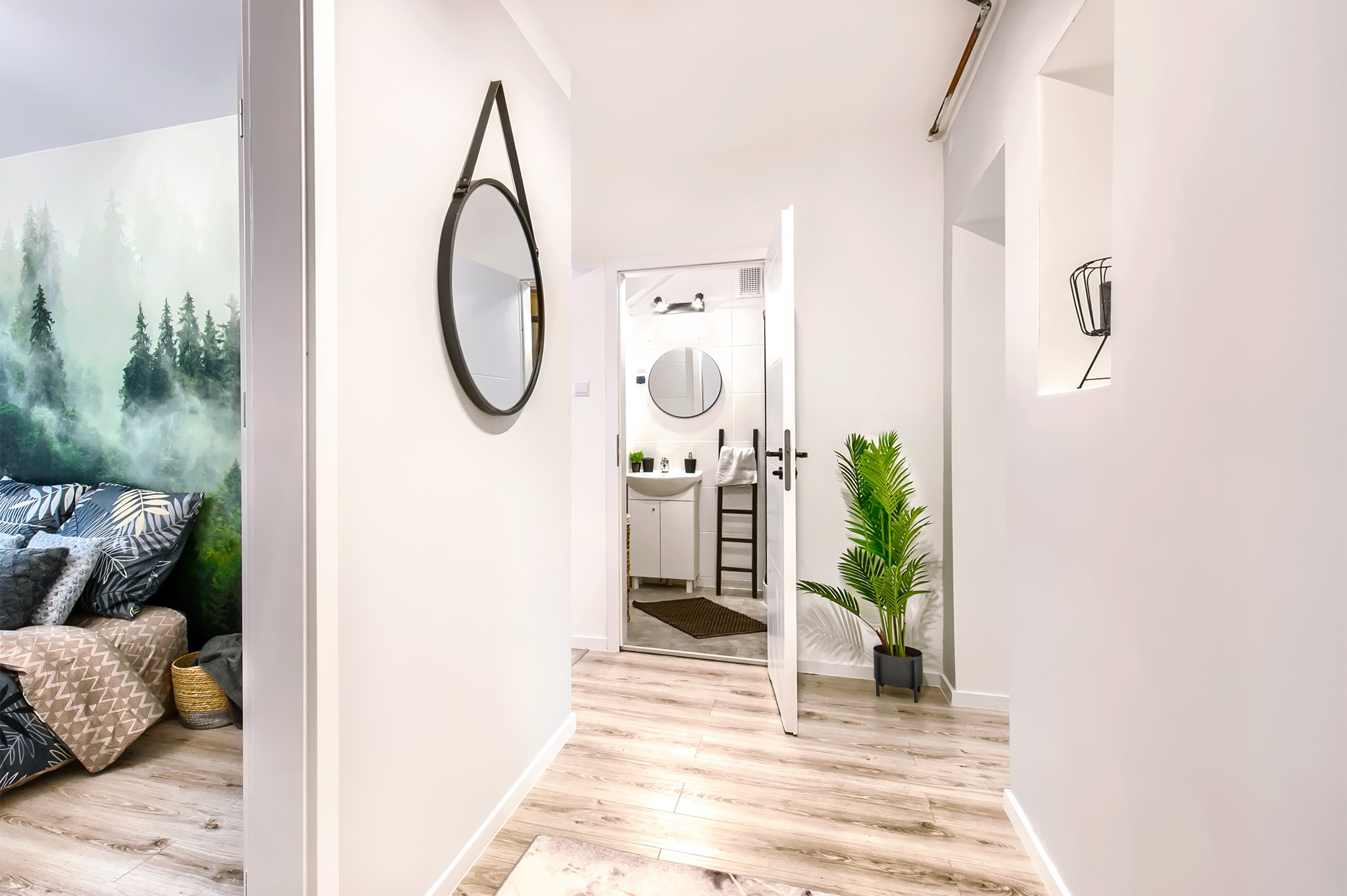 Wnętrze mieszkania po liftingu: widok na korytarz z lustrem w skórzanej ramie, otwarte drzwi do łazienki z czarną drabinką na ręczniki i widok na sypialnię z fototapetą leśną, jasne drewniane...