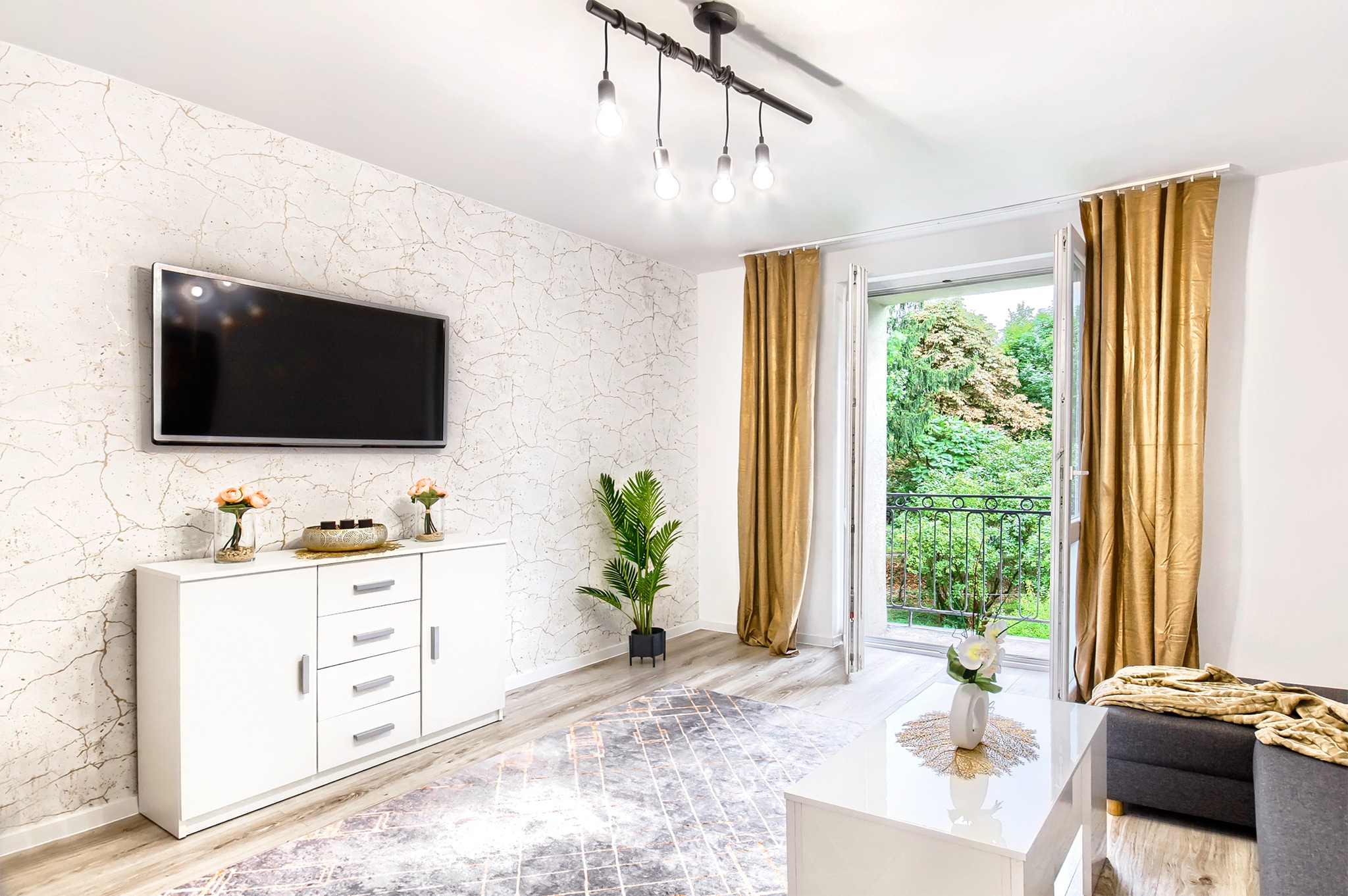 Jasny salon po liftingu z telewizorem na ścianie o wzorze marmuru, białą komodą, rośliną doniczkową, i wyjściem na balkon z zielonym widokiem.