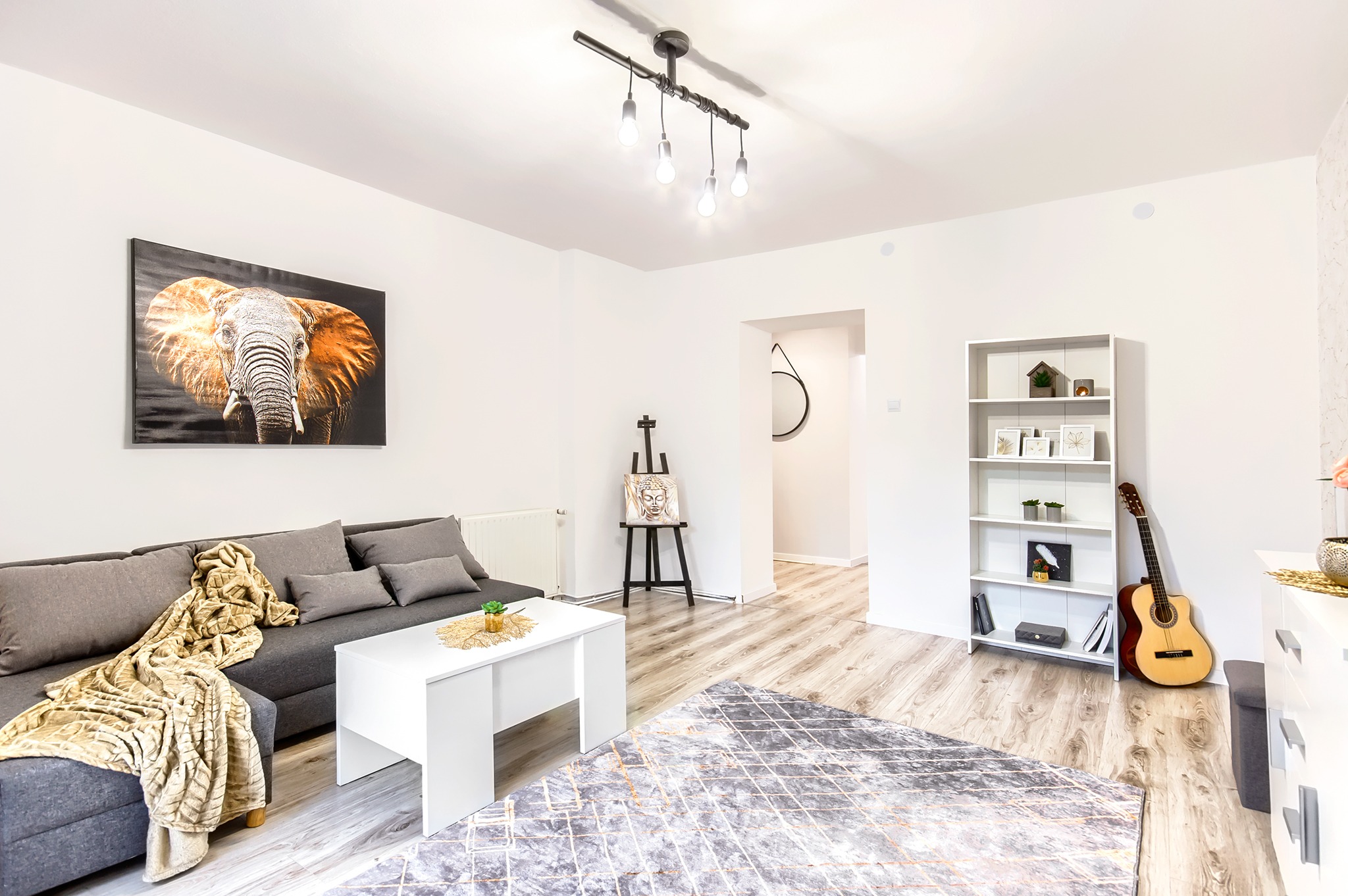 Jasny salon po liftingu z szarą sofą, białym stolikiem kawowym, obrazem słonia na ścianie i gitarą opartą o regał.