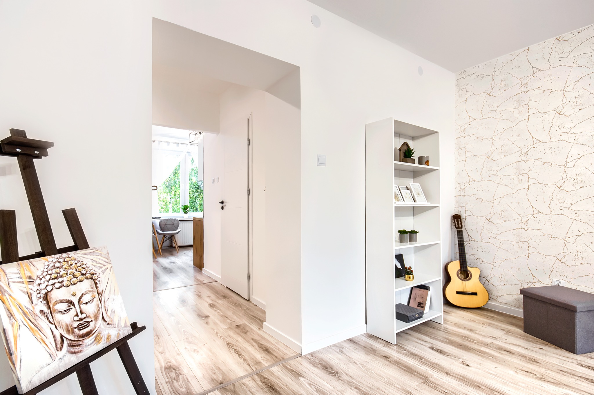 Jasne wnętrze mieszkania po liftingu: parkiet w odcieniach drewna, białe ściany, obraz Buddy na sztaludze, gitara oparta o regał na tle tapety z motywem marmuru.