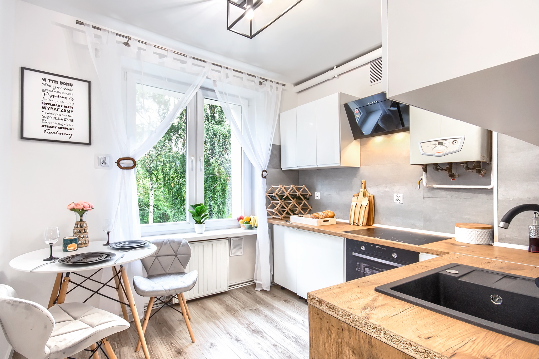 Jasna kuchnia po liftingu i home stagingu: drewniany blat, szara ściana, białe szafki, okrągły stół z szarymi krzesłami, widok na zieleń za oknem, dekoracyjny napis na ścianie.