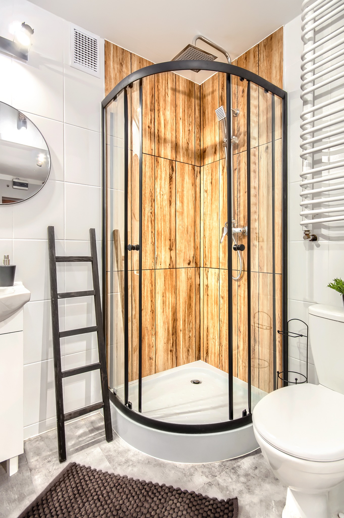 Nowoczesna łazienka z kabiną prysznicową z czarnymi profilami i drewnopodobną okładziną, drabina dekoracyjna, biała armatura i szara podłoga.