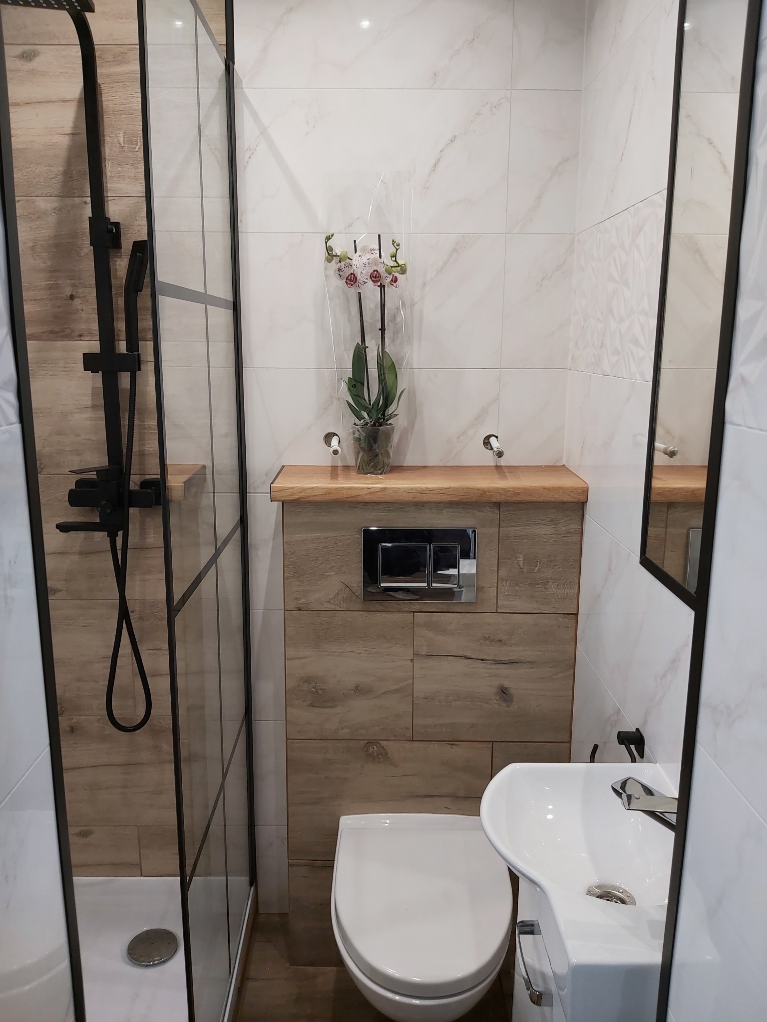 Zakończona aranżacja małej łazienki z kabiną prysznicową typu walk-in z czarną armaturą, wiszącą toaletą, umywalką i drewnianym blatem nad stelażem, ozdobiona kwiatem orchidei.