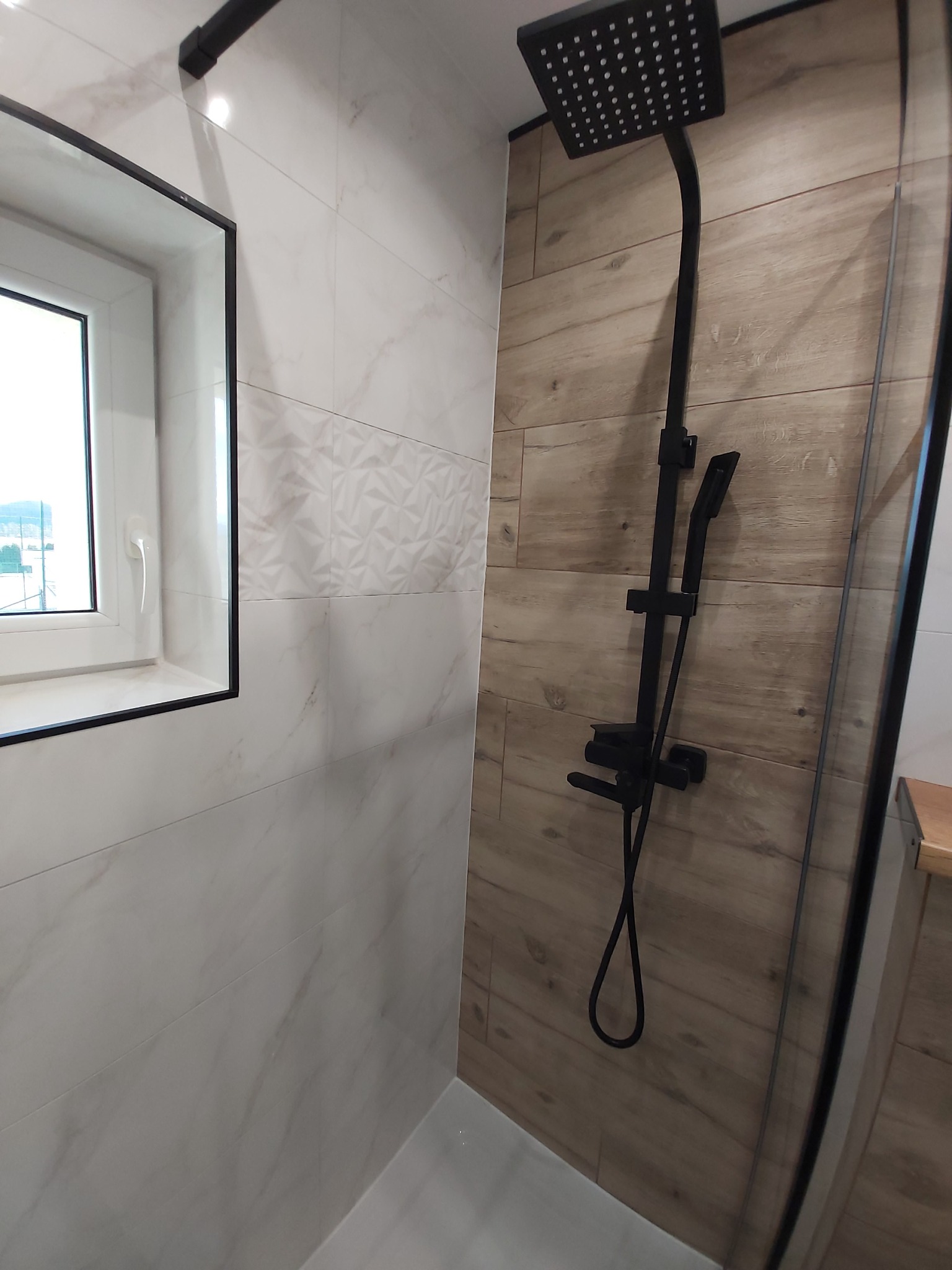 Nowoczesna kabina prysznicowa z czarną armaturą, wykończona płytkami imitującymi drewno i marmur, z oknem w czarnej ramie.