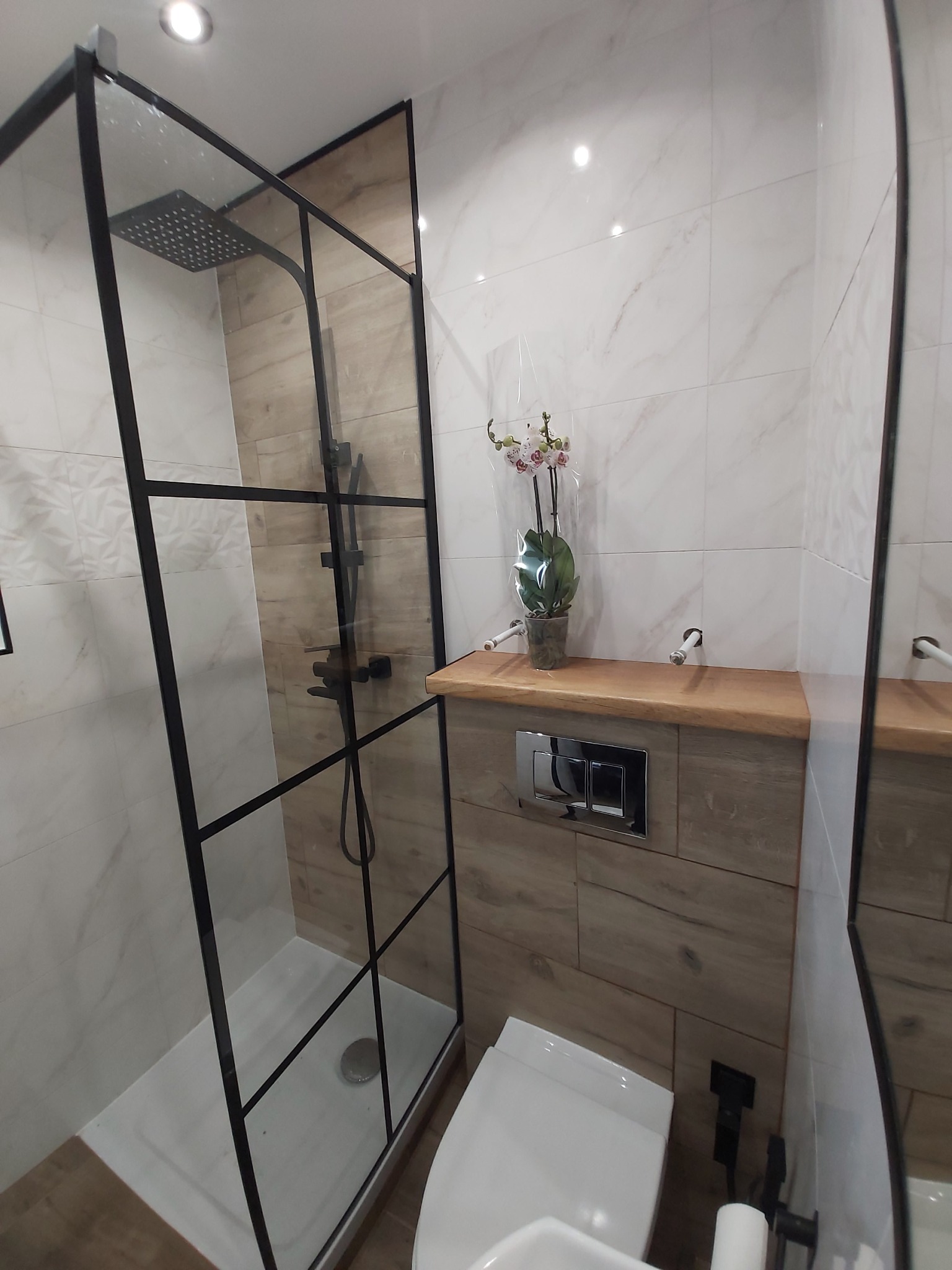 Nowoczesna łazienka po remoncie z kabiną prysznicową typu walk-in z czarnymi profilami, wiszącą toaletą, drewnianym blatem i marmurowymi płytkami ściennymi, ozdobiona storczykiem w doniczce.