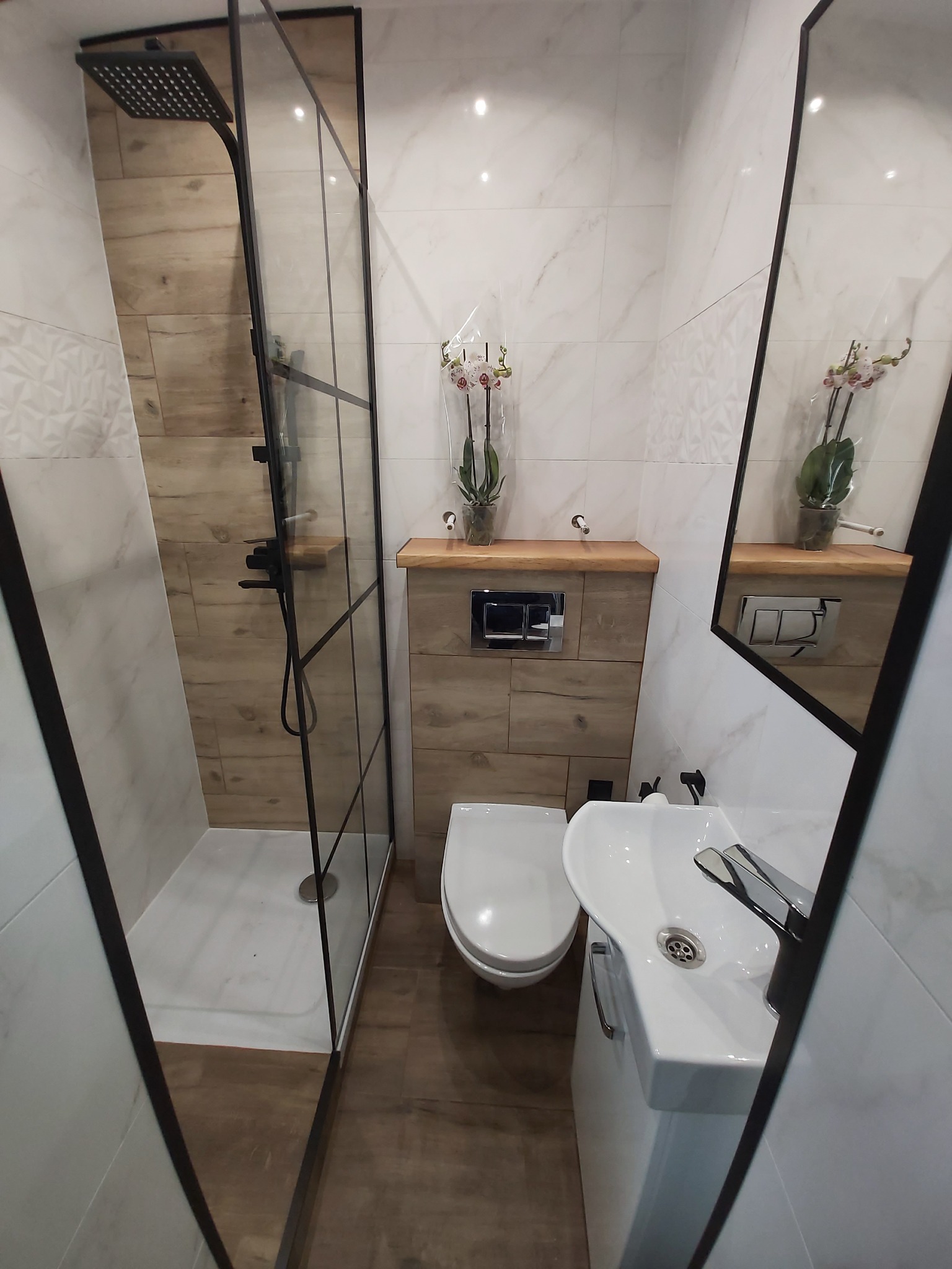 Nowoczesna łazienka z kabiną prysznicową typu walk-in z czarnymi ramami, wiszącą toaletą, umywalką na szafce i lustrem, wykończona płytkami imitującymi drewno i białymi płytkami z delikatnym wzorem.