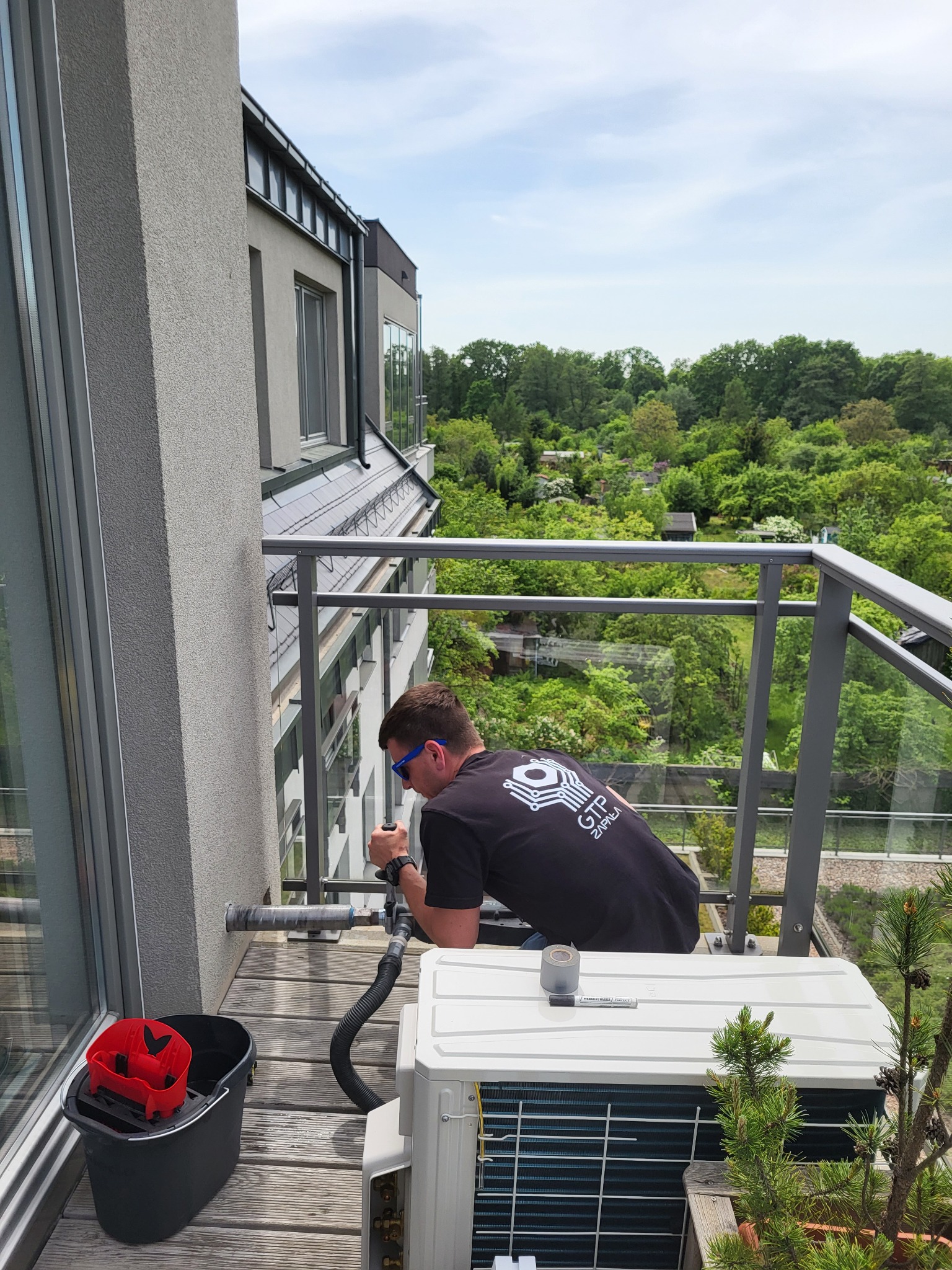 Monter w okularach wierci otwór wiertnicą na sucho w ścianie na balkonie, podłączoną do odkurzacza, obok stoi wiadro z akcesoriami i jednostka zewnętrzna klimatyzatora.