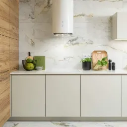 Jasna kuchnia z marmurowymi płytkami na ścianie i podłodze, drewnianymi szafkami i cylindrycznym okapem nad płytą grzewczą, uzupełniona zielonymi jabłkami, ziołami w doniczce i książką kucharską.