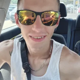 Autoportret uśmiechniętego mężczyzny w okularach przeciwsłonecznych, siedzącego w samochodzie w Nowej Rudzie, z widocznym odbiciem kierowcy i wnętrza auta w szkłach.