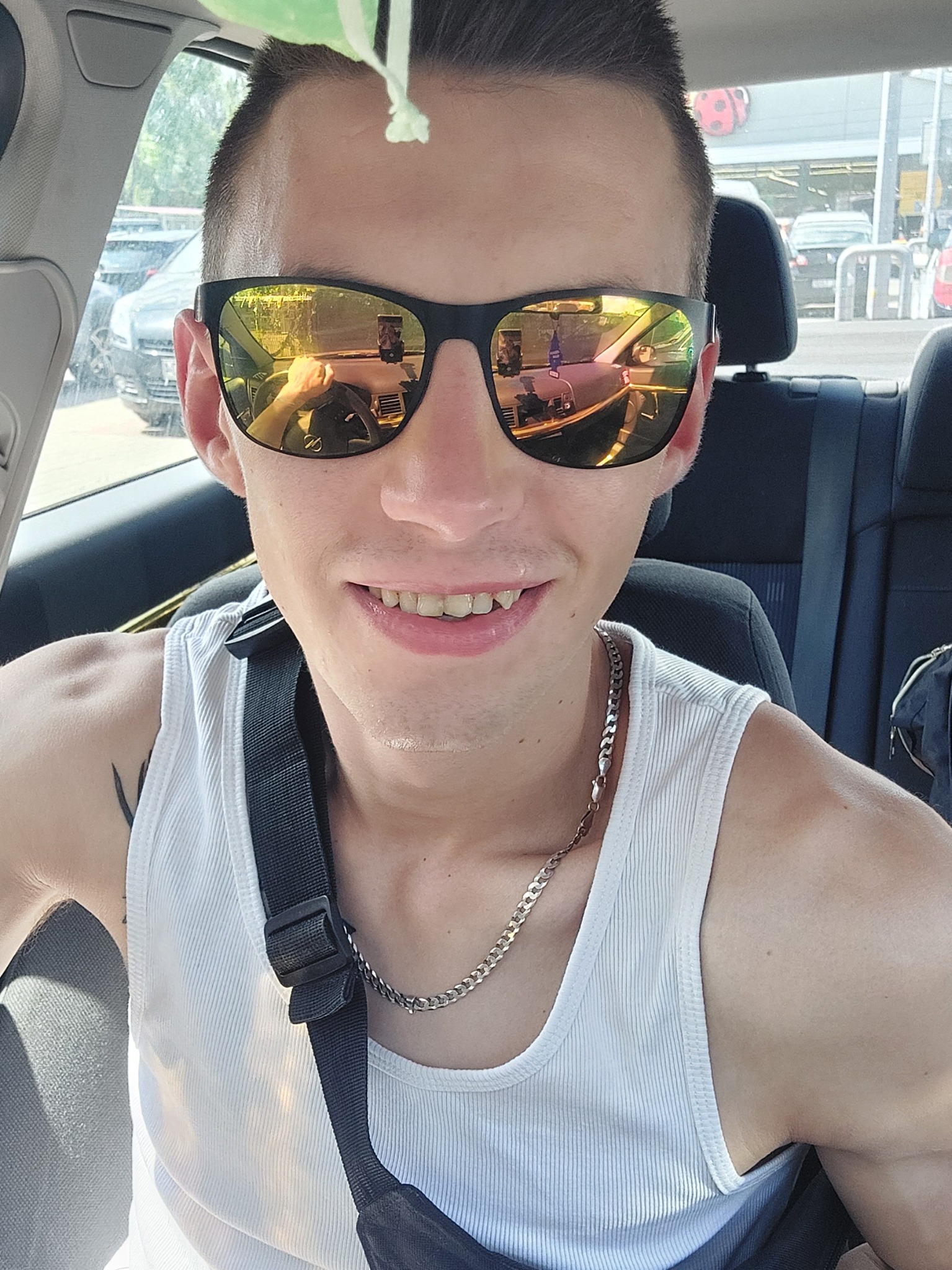 Autoportret uśmiechniętego mężczyzny w okularach przeciwsłonecznych, siedzącego w samochodzie w Nowej Rudzie, z widocznym odbiciem kierowcy i wnętrza auta w szkłach.