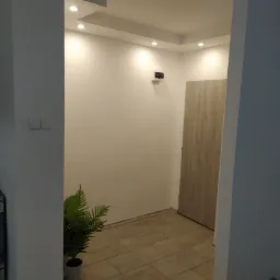 Wnętrze mieszkania po remoncie: białe ściany z oświetleniem punktowym, jasna podłoga z płytek, drzwi wewnętrzne, roślina doniczkowa i fragment kuchenki z ręcznikiem w tropikalne liście.