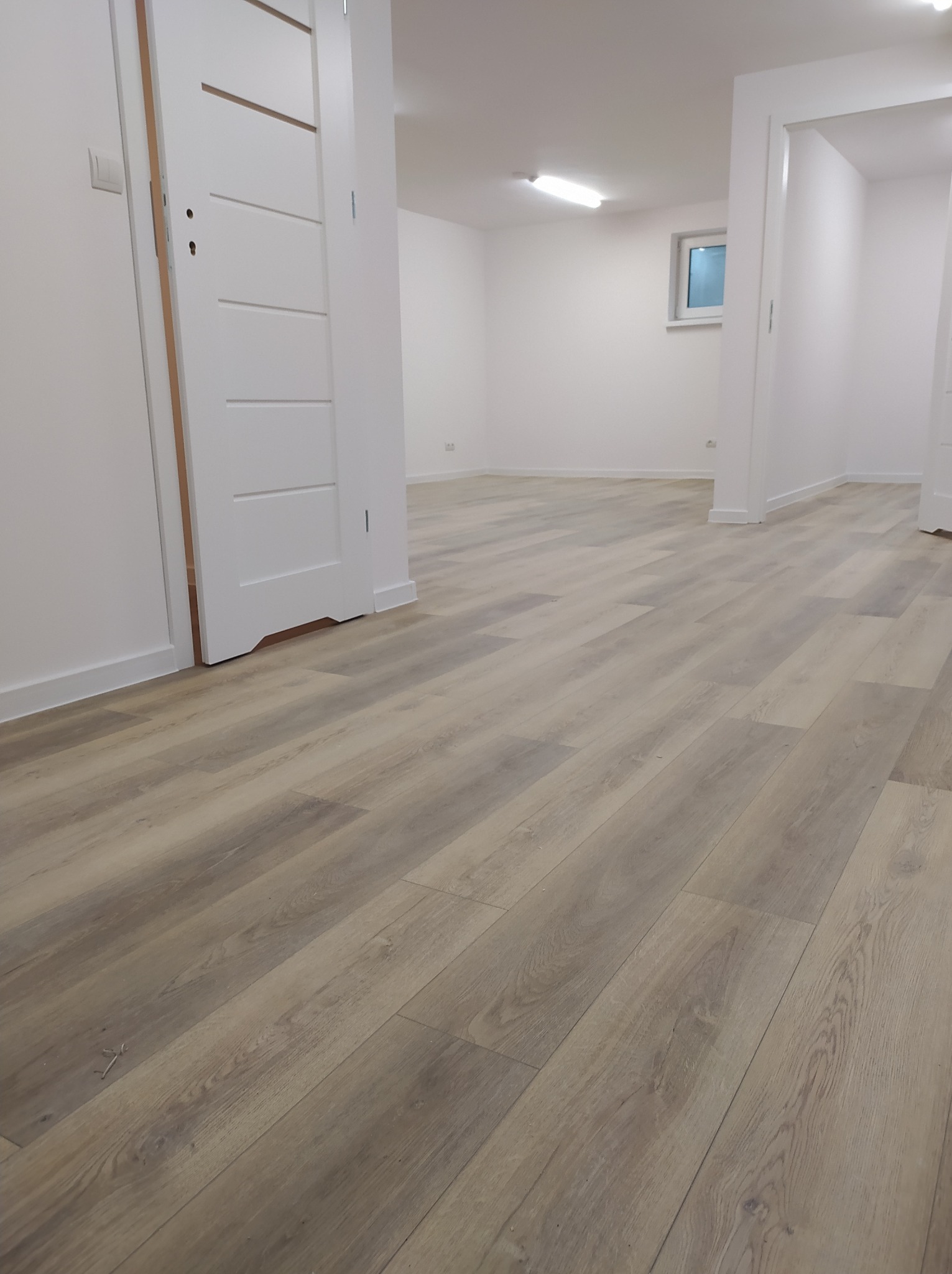 Ułożone panele podłogowe w jasnym odcieniu drewna w nowym, pustym pomieszczeniu z białymi ścianami i drzwiami.