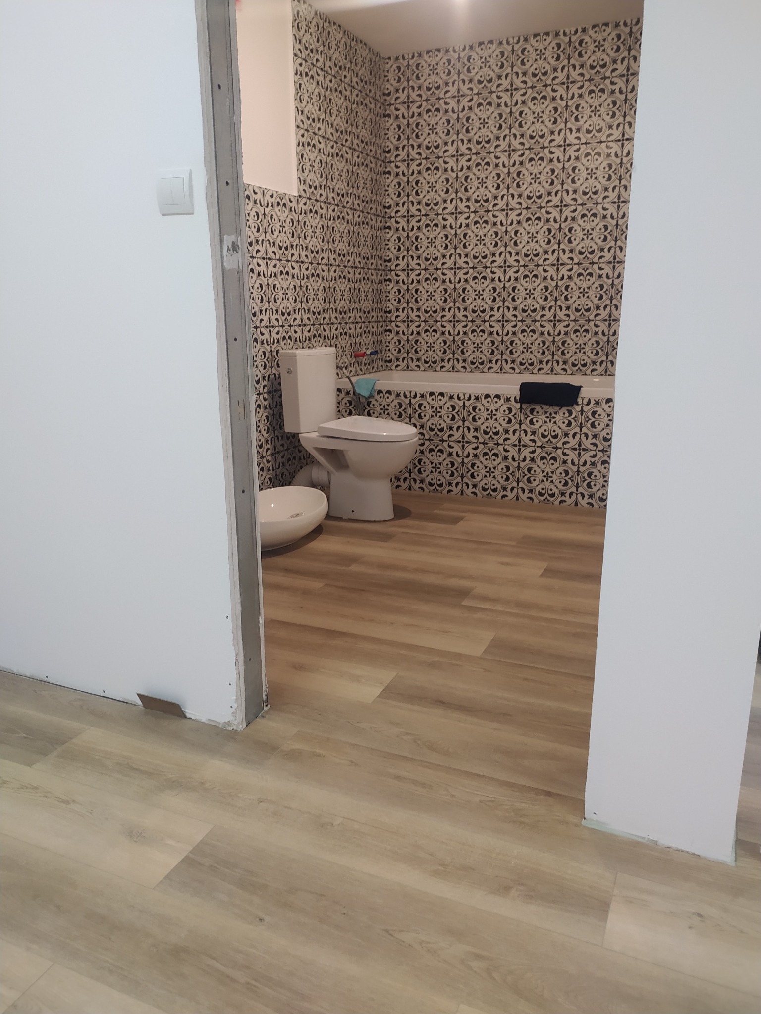 Wykończona łazienka z wzorzystymi płytkami na ścianie nad wanną, umywalką i toaletą, oraz panelami podłogowymi o jasnym odcieniu drewna, widoczna zza niedokończonego ościeża drzwiowego.