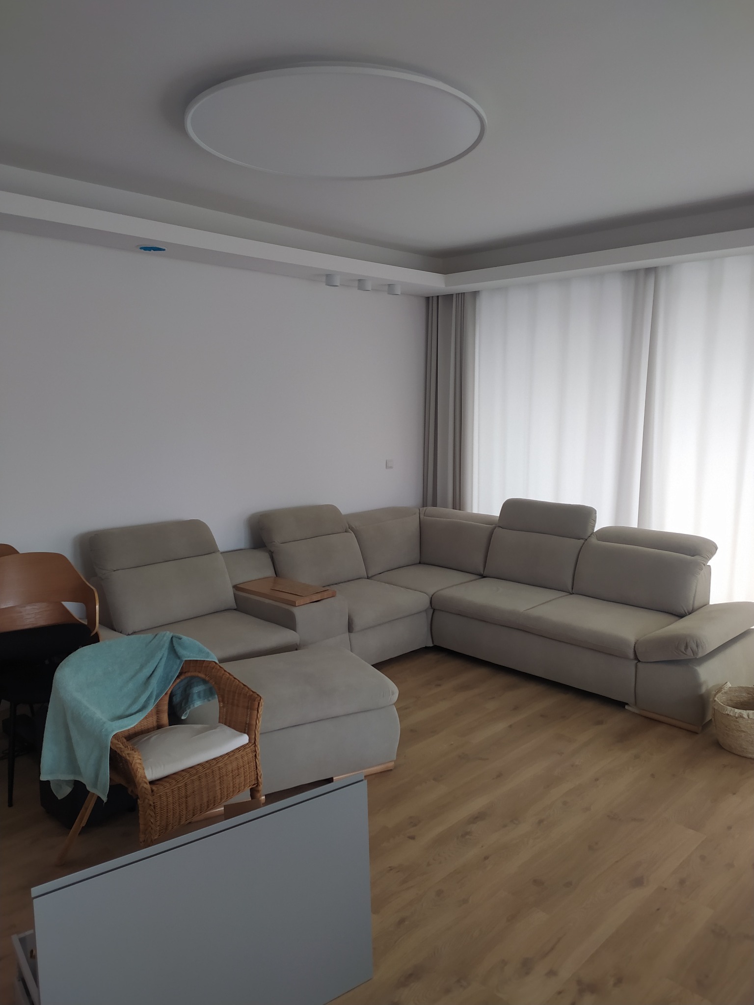 Jasny salon z narożną sofą z regulowanymi zagłówkami, drewnianą podłogą i rattanowym fotelem z turkusowym pledem