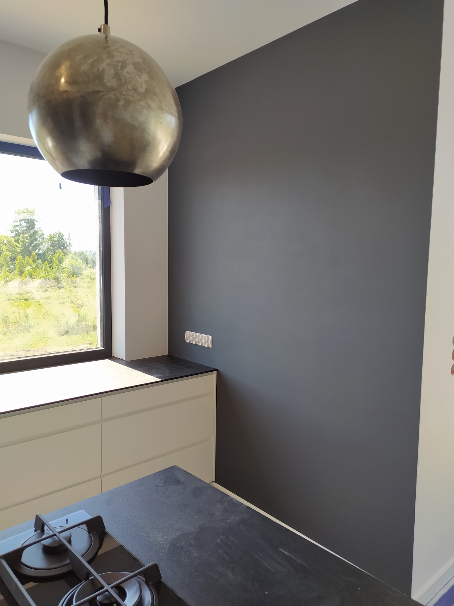 Wnętrze kuchni z szarą ścianą, okrągłą lampą wiszącą i fragmentem blatu kuchennego z płytą gazową.