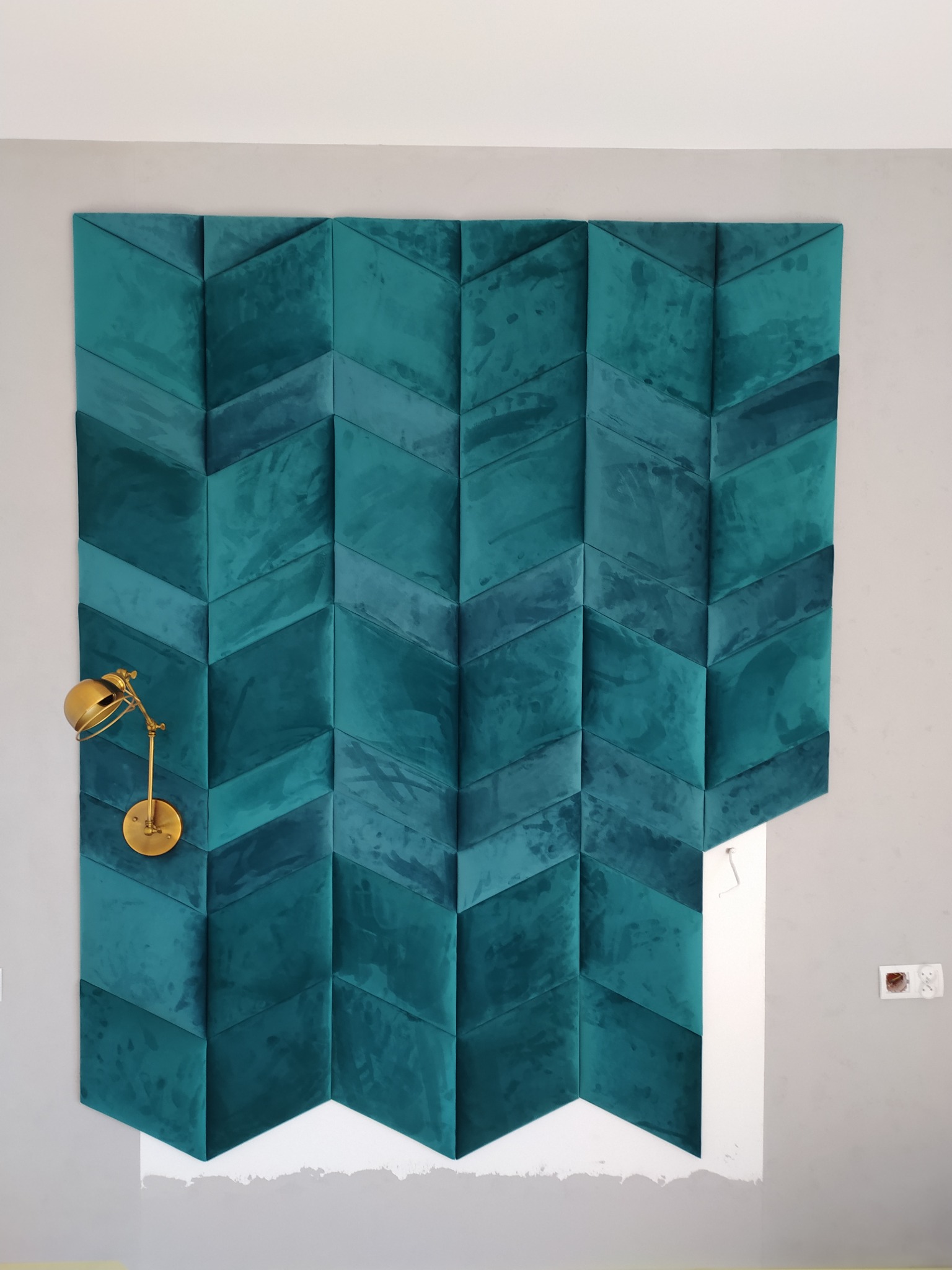 Turkusowy panel ścienny z geometrycznym wzorem w jodełkę, oświetlony złotą lampką, w trakcie montażu widoczne niedomalowane fragmenty ściany.