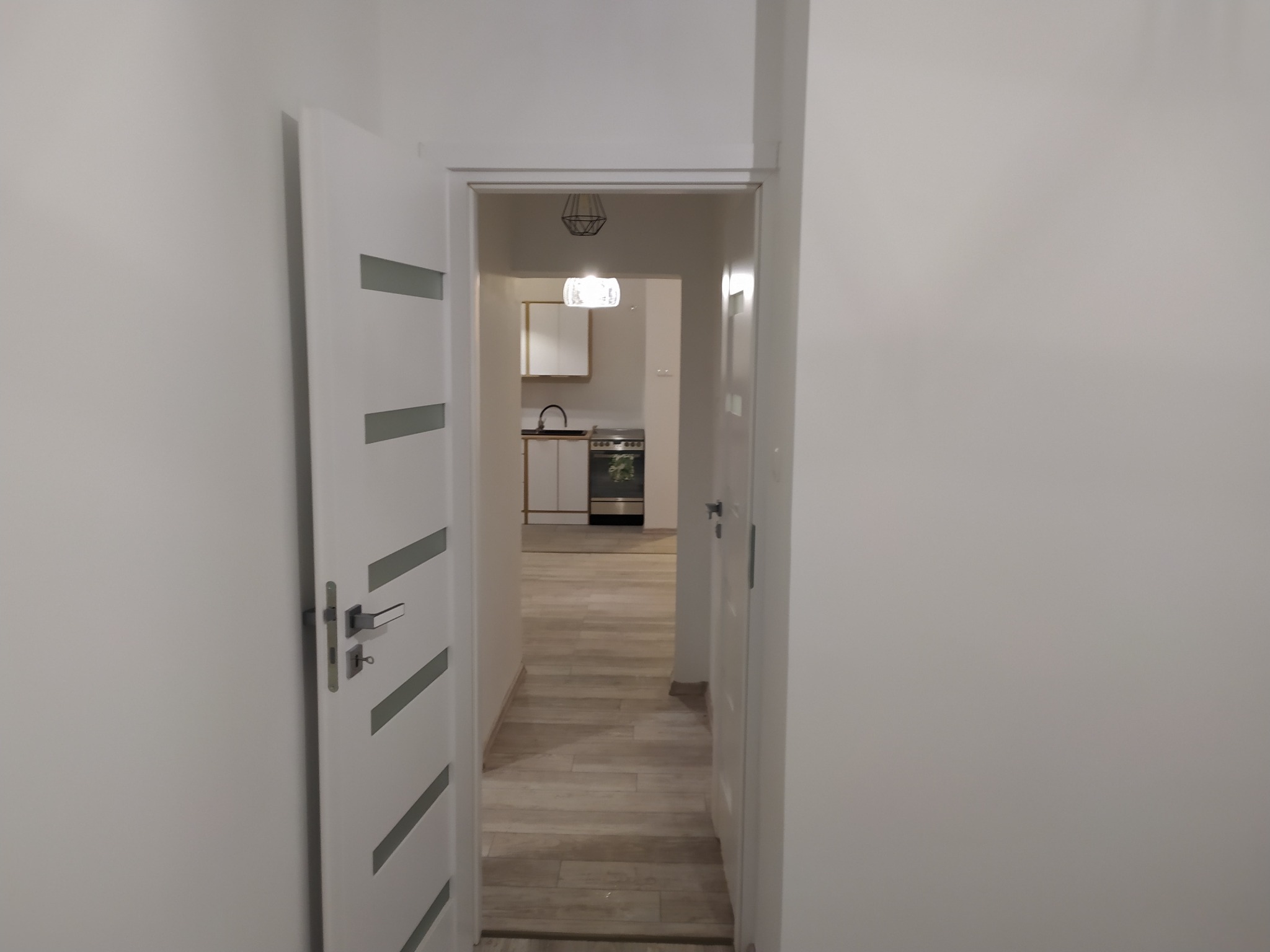 Widok przez otwarte drzwi na odnowione wnętrze mieszkania z jasnymi ścianami i panelami podłogowymi, w oddali widoczna kuchnia z białymi szafkami i nowoczesną kuchenką.