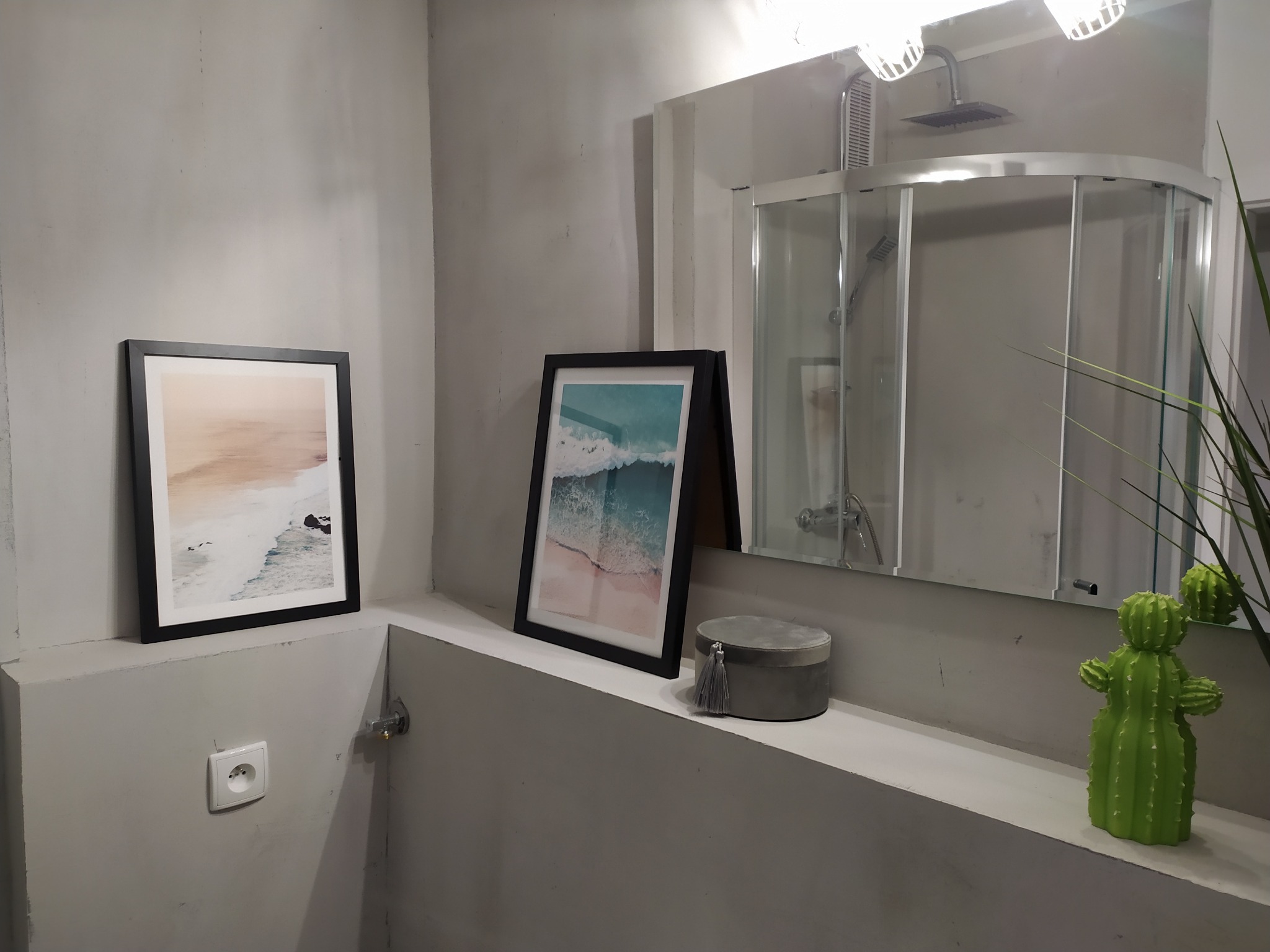 Szara łazienka z kabiną prysznicową, dekoracyjnymi obrazami w czarnych ramkach, ceramicznym kaktusem i pudełkiem z frędzlami.