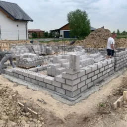 Wznoszenie ścian fundamentowych z bloczków betonowych na budowie domu jednorodzinnego; widoczni dwaj pracownicy, jeden z taczką, drugi przy murze; w tle dom w stanie surowym otwartym i sterta ziemi.