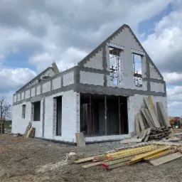 Surowy stan domu z białych bloczków, widoczne zbrojenia okien, drewniane stemple podtrzymujące nadproże, składowane deski i materiały budowlane na placu budowy w pochmurny dzień.