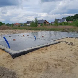 Świeżo wylane, mokre fundamenty z wystającymi rurami drenażowymi na piaszczystym terenie budowy, w tle domy i pochmurne niebo.