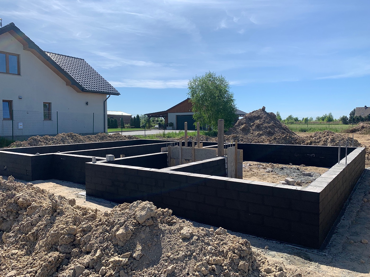 Wykopane i częściowo wylane fundamenty pod dom jednorodzinny z widocznymi zbrojeniami i izolacją pionową, w tle dom w trakcie budowy i sterta ziemi.