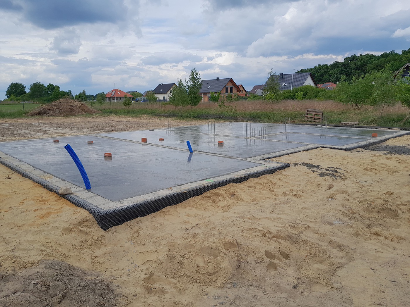 Świeżo wylane, mokre fundamenty z wystającymi rurami drenażowymi na piaszczystym terenie budowy, w tle domy i pochmurne niebo.