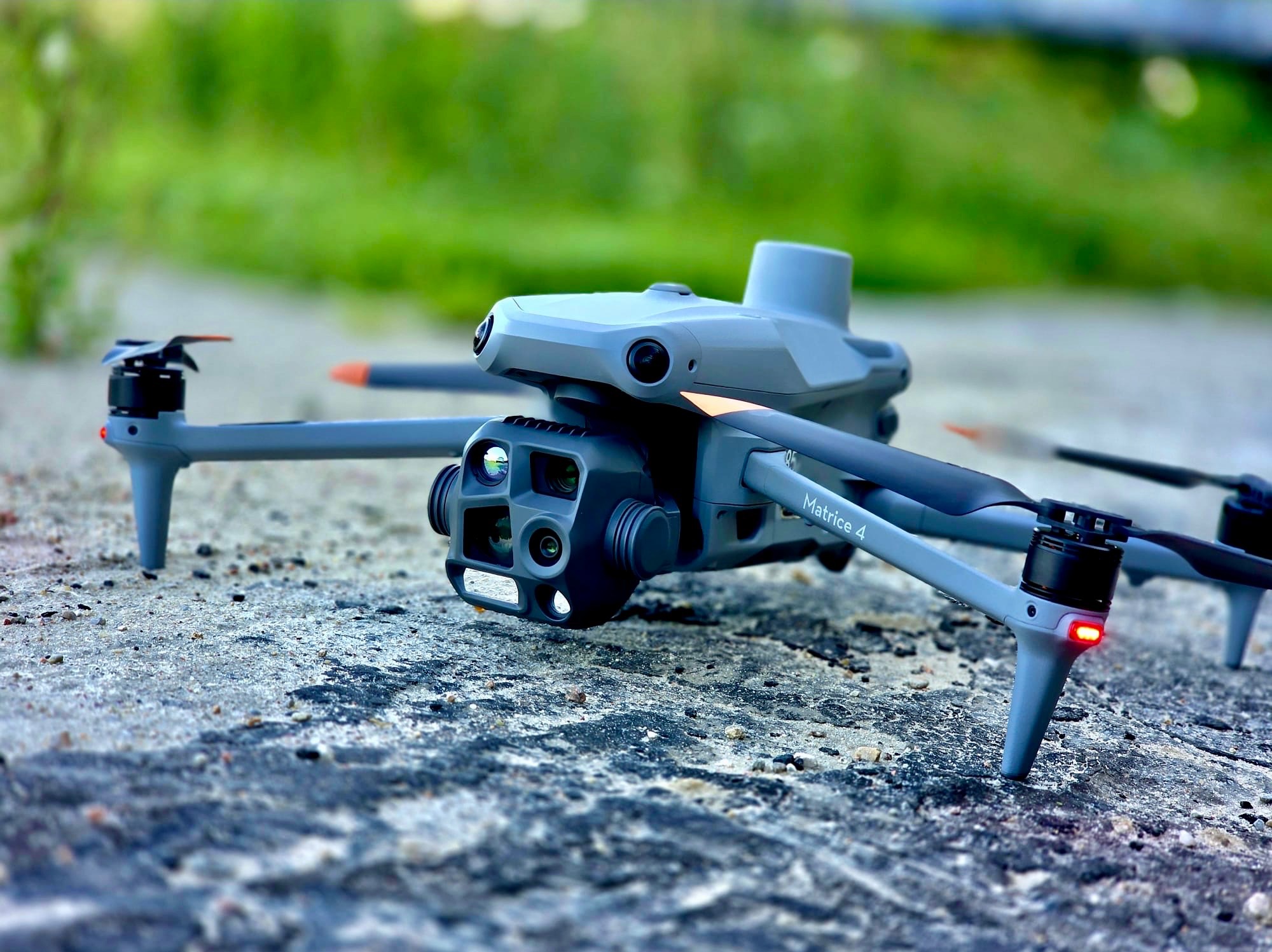 Szary dron Matrice 4 leżący na szorstkiej, jasnej powierzchni z zielonym, rozmytym tłem. Widoczne kamery i czujniki drona.