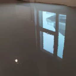 Świeżo wylany, szary mikrocement na podłodze w jasnym pokoju z grzejnikiem i oknem, w którym odbija się budynek w budowie.