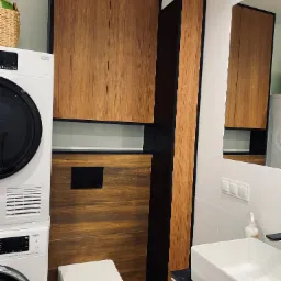 Nowoczesna łazienka z drewnianymi szafkami, zabudową WC, pralką i umywalką nablatową. Szafki z ciemnymi obramowaniami, białe płytki na ścianach. W lustrze odbija się fragment innej szafki. W rogu...
