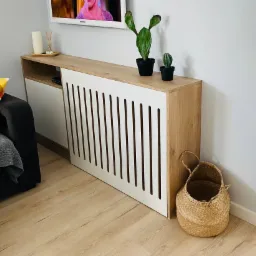 Biało-drewniana zabudowa grzejnika z pionowymi szczelinami, na niej dwa kaktusy w czarnych doniczkach, obok koszyk z trawy morskiej, w tle fragment szarej sofy z kolorową poduszką.