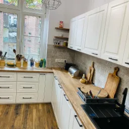 Jasna kuchnia z drewnianym blatem, białymi szafkami z czarnymi uchwytami, wzorzystymi płytkami nad blatem i drewnianą podłogą. Na blacie widoczne akcesoria kuchenne, deski do krojenia i suszarka...