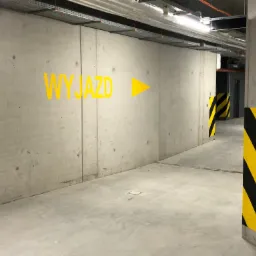 Betonowa posadzka i ściany w garażu podziemnym z żółtym napisem 'WYJAZD' i strzałką, słupy oznaczone czarno-żółtymi pasami.
