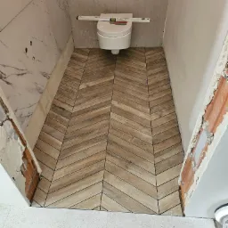 Ukończona podłoga w małej łazience z drewnopodobnych płytek ułożonych w jodełkę, częściowo wykończone ściany z białego marmuru i szarej płyty gipsowej, poziomica na sedesie