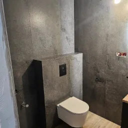 Zawieszona miska WC na tle szarych, kamiennych płytek w nowoczesnej łazience, widoczne drewnopodobne panele podłogowe i czarne elementy armatury.