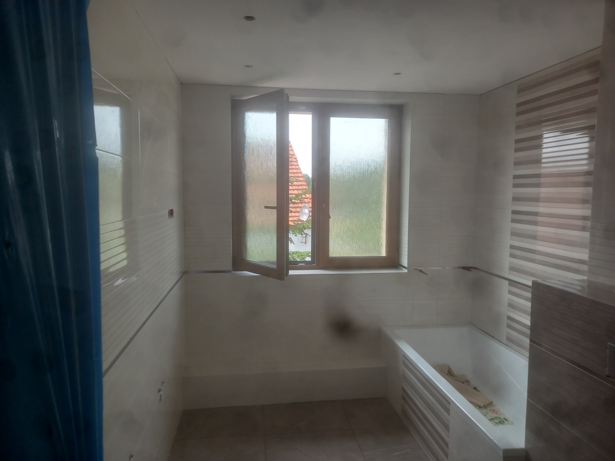 Wykończona łazienka z biało-szarymi płytkami, otwartym oknem z widokiem na dach i zieloną roślinność, oraz wanną z ręcznikiem w środku.