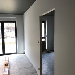 Łukasz, kafle, remonty, wykonczenia. - Wnętrze w trakcie remontu: szare płytki podłogowe, świeżo pomalowane ściany na szaro, ciemnoszary sufit, kartony z materiałami budowlanymi ustawione wzdłuż ściany, widok przez drzwi do kolejnego...