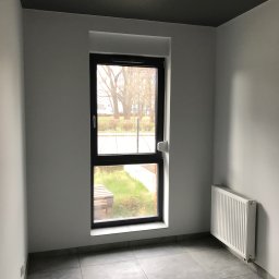 Łukasz, kafle, remonty, wykonczenia. - Pomalowane na szaro wnętrze pokoju z grzejnikiem, szarymi płytkami podłogowymi i oknem z widokiem na zewnątrz.