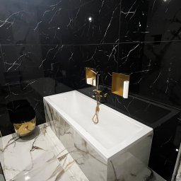 Łukasz, kafle, remonty, wykonczenia. - Nowoczesna łazienka z czarnymi płytkami imitującymi marmur na ścianach, białą prostokątną wanną obudowaną marmurem, złotą armaturą i czarno-złotą toaletą.