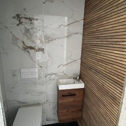 Łukasz, kafle, remonty, wykonczenia. - Nowoczesna łazienka z marmurowymi płytkami ściennymi, drewnianą boazerią, wiszącą toaletą i umywalką z szafką z ciemnego drewna na podłodze z ciemnego marmuru.
