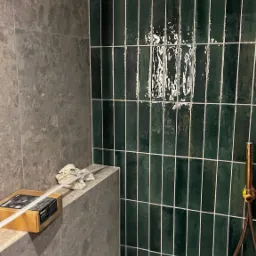 Fragment łazienki z ciemnozielonymi, błyszczącymi, pionowo ułożonymi płytkami typu 'cegiełka' z jasną fugą, szarym marmurem na sąsiedniej ścianie i złotym prysznicem, z pudełkiem narzędzi na półce.