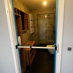 Łukasz, kafle, remonty, wykonczenia. - Zabezpieczone wejście do łazienki z czarną armaturą i drewnianymi meblami, widoczne narzędzie do zabezpieczania futryny, podłoga z ciemnego marmuru.