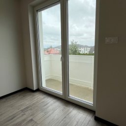 Łukasz, kafle, remonty, wykonczenia. - Nowoczesne wnętrze z drewnianą podłogą i dużym oknem balkonowym, przez które widać balkon z białą balustradą i zabudowę miejską w tle.