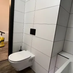 Nowoczesna łazienka z białymi, prostokątnymi płytkami i drewnopodobną podłogą. Wisząca toaleta i fragment wanny. Widoczny odkurzacz w tle. Minimalistyczny design.