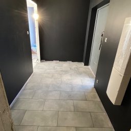 Łukasz, kafle, remonty, wykonczenia. - Wykończony korytarz z szarymi płytkami podłogowymi i czarnymi ścianami. Widoczne drzwi i wisząca żarówka. Minimalistyczny design.