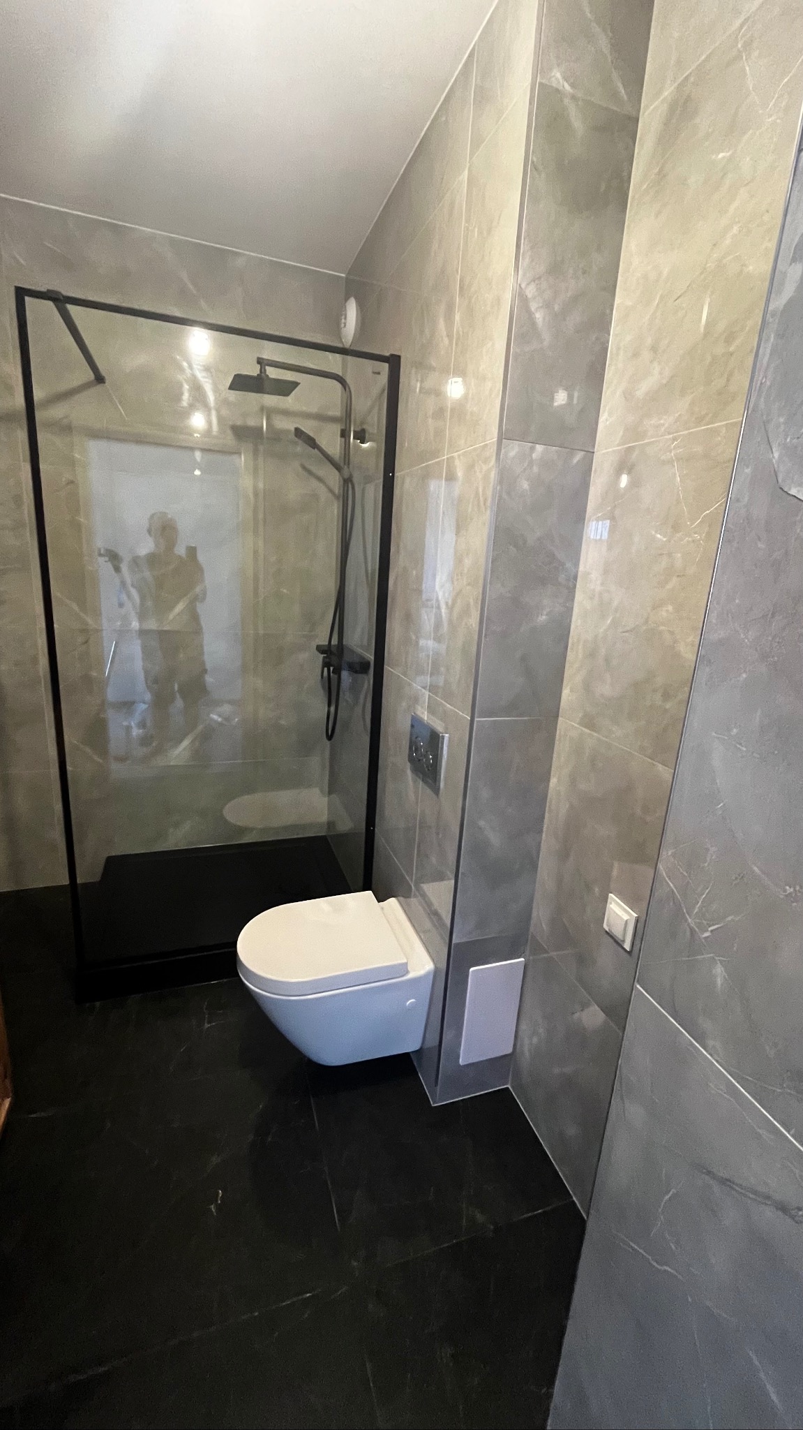 Nowoczesna łazienka z czarną kabiną prysznicową typu walk-in, podwieszaną toaletą i szarymi płytkami imitującymi marmur na ścianach i czarną marmurową podłogą.