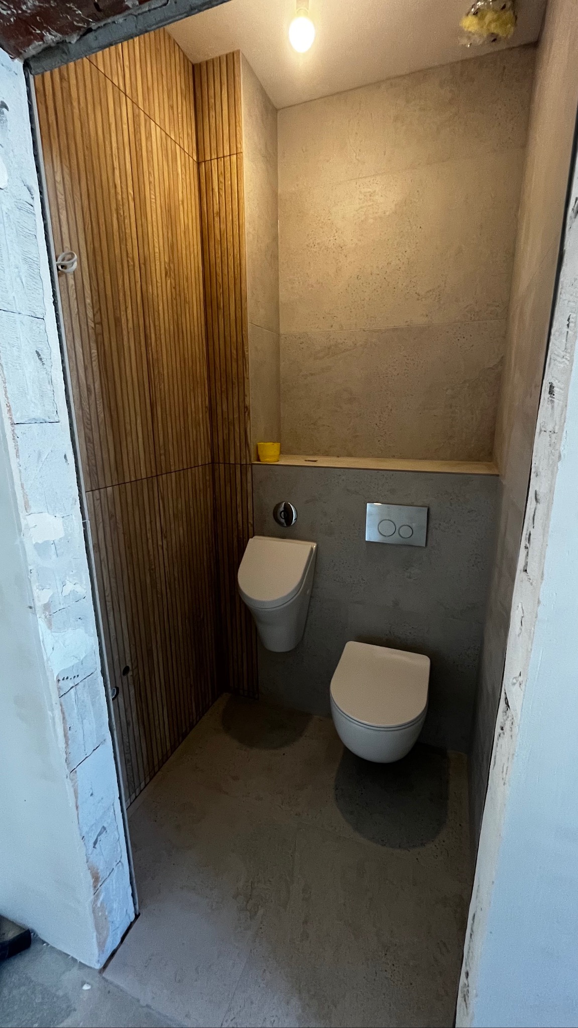 Wnęka toaletowa z drewnianą boazerią na jednej ścianie, wiszącą muszlą klozetową i pisuarą, szare płytki na ścianach i podłodze, widoczne surowe wykończenie przy drzwiach.