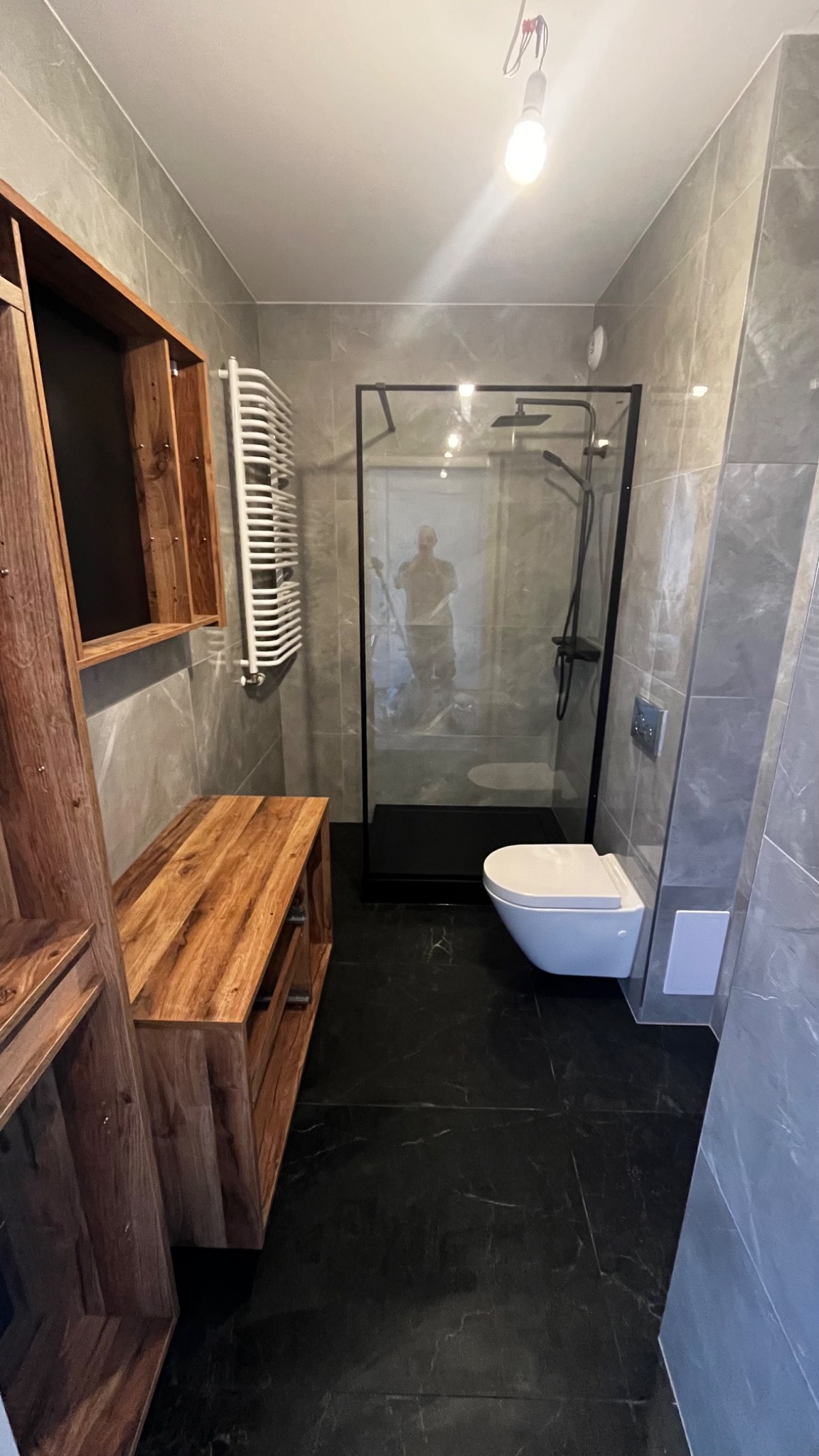 Nowoczesna łazienka z szarymi płytkami imitującymi marmur, czarną podłogą i kabiną prysznicową typu walk-in z czarnymi ramami, drewnianą szafką z otwartymi półkami i grzejnikiem drabinkowym.